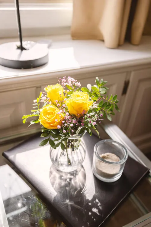 Ein Strauß gelber Rosen und ein Glas Wasser auf einem Tisch mit einem schwarzen Buch.