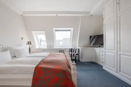 Elegantes Hotelzimmer mit Doppelbett, modernem Fernseher, gestreiftem Stuhl und natürlichem Licht durch schrägstehende Fenster.